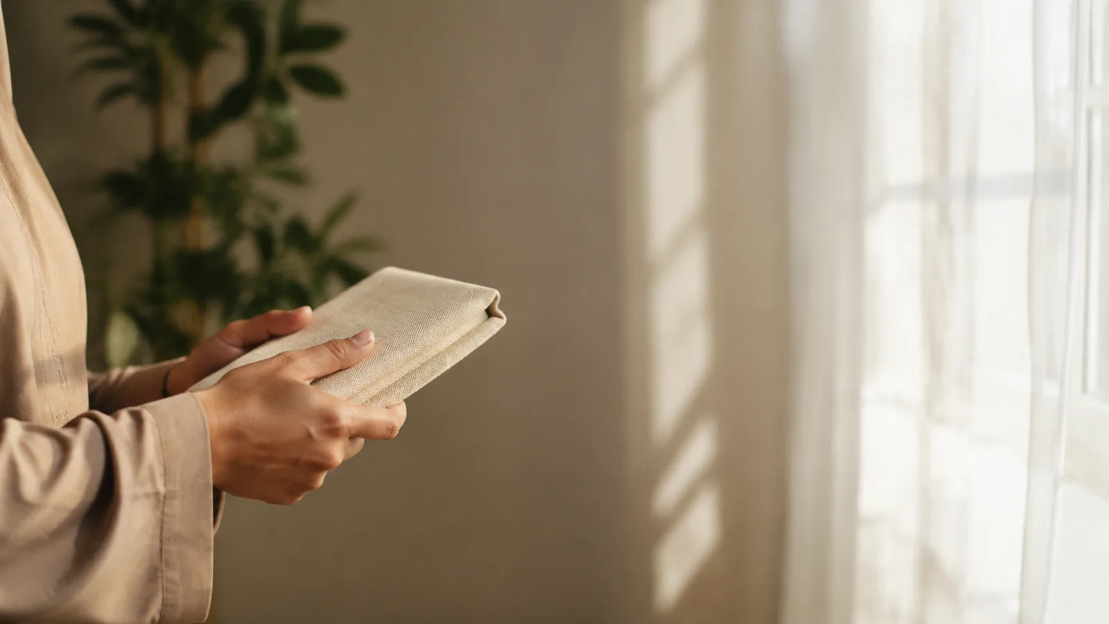 A person's hands holding a closed Quran at a window with daylight falling across it
