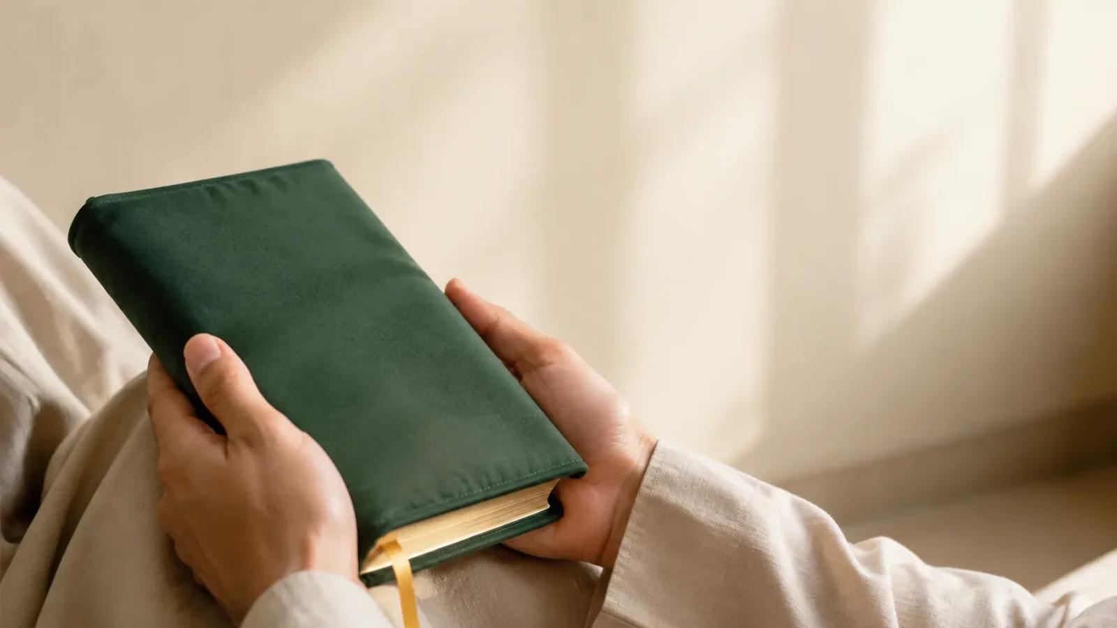 Hands holding a closed green Quran with a ribbon bookmark in a quiet, softly lit room